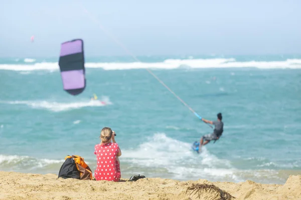 Mädchen sitzt am Sandstrand und beobachtet Kitesurfer im Meer. — Stockfoto