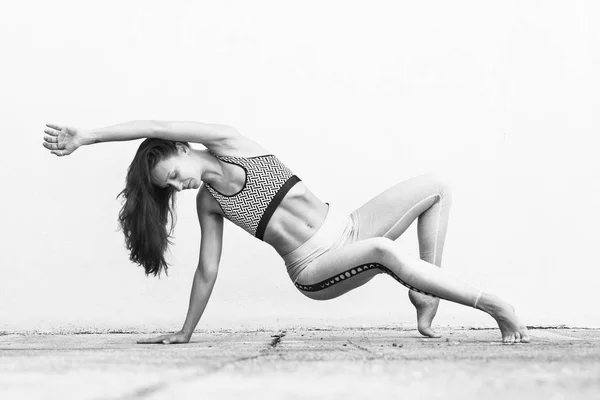Fit sporty active girl in fashion sportswear doing yoga fitness exercise in front of gray wall, outdoor sports, urban style. Black and white photo. — Stock Photo, Image