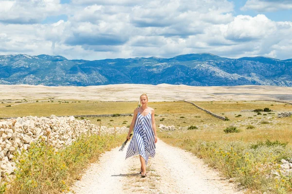 Mujer joven caucásica en vestido de verano con ramo de flores de lavanda mientras camina al aire libre a través del paisaje seco rocoso mediterráneo de la costa croata en la isla de Pag en verano —  Fotos de Stock
