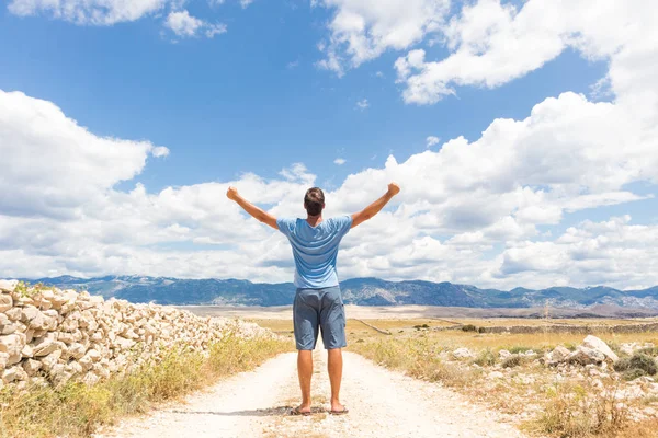 Rear view of casual sporty man standing on a dirt country road rising hands up to the clouds on a blue summer sky. Freedom and travel adventure concept. — Stock Photo, Image