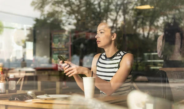 Thoughtful caucasian woman holding mobile phone while looking through the coffee shop window during coffee break.