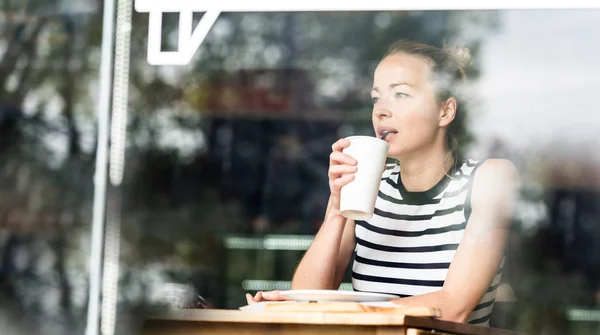 Jovem caucasiana sentada sozinha em uma cafetaria pensativa apoiada na mão, olhando pela janela — Fotografia de Stock