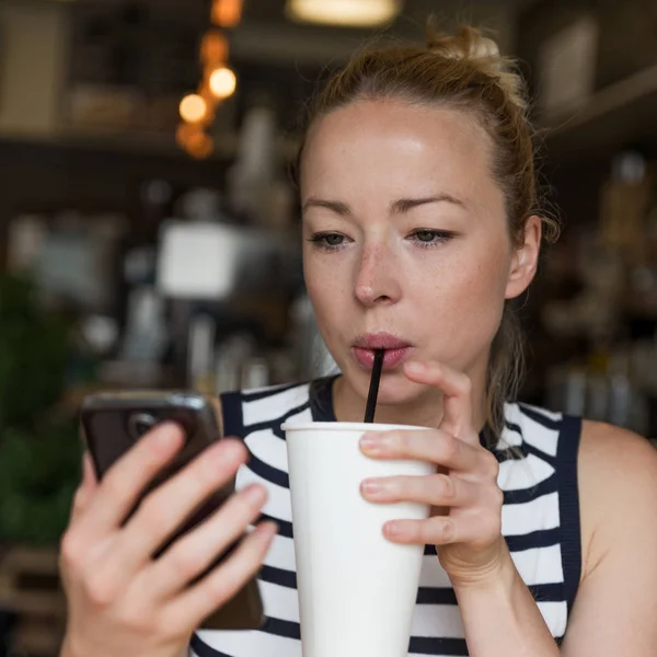 Tankeväckande kvinna läser nyheter på mobiltelefon samtidigt smuttar kaffe i caféet. — Stockfoto