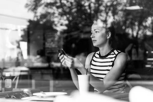 Kahve molasında kahve dükkanının camından bakarken cep telefonu tutan düşünceli beyaz kadın.. — Stok fotoğraf