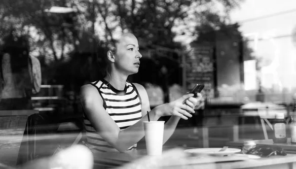 Στοχαστική καυκάσιος γυναίκα κρατώντας το κινητό τηλέφωνο, ενώ κοιτάζοντας μέσα από το παράθυρο της καφετέριας κατά τη διάρκεια διάλειμμα καφέ. — Φωτογραφία Αρχείου