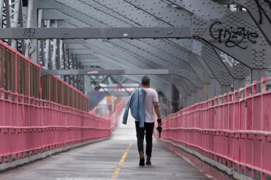 Williamsburg Köprüsü, Brooklyn, New York'ta yürürken omzunun üzerinde kot ceket taşıyan tanınmaz şık genç adam Arka görünümü.
