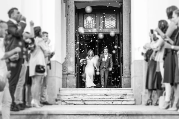 Recém-casados saindo da igreja após a cerimônia de casamento, família e amigos comemorando seu amor com o chuveiro de bolhas de sabão, costume minando banho de arroz tradicional — Fotografia de Stock