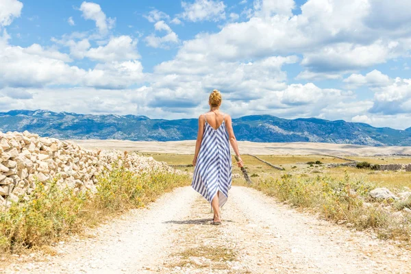 Задний вид женщины в летнем платье, держащей букет лавандовых цветов во время прогулки на открытом воздухе через сухой скалистый средиземноморский хорватский берег Lanscape на острове Паг летом — стоковое фото