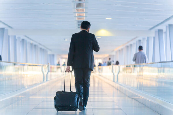 Businessman walking and wheeling a trolley suitcase at the lobby, talking on a mobile phone. Business travel concept.