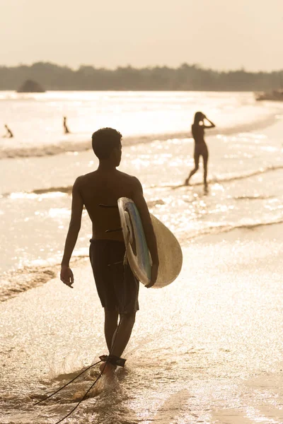 Человек, идущий по тропическому пляжу Мидигамы, Шри-Ланка на закате с доской для серфинга в руках . — стоковое фото