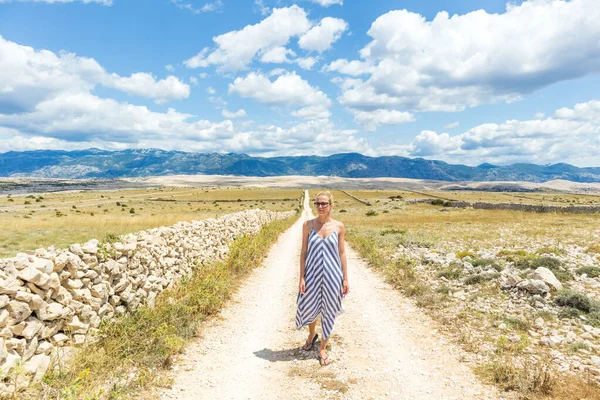 Mujer joven caucásica en vestido de verano con ramo de flores de lavanda mientras camina al aire libre a través del paisaje seco rocoso mediterráneo de la costa croata en la isla de Pag en verano —  Fotos de Stock