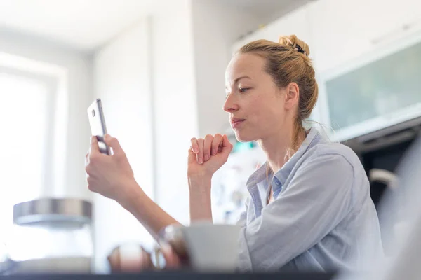 Młoda zadowolona kobieta w domu kuchnia za pomocą aplikacji mediów społecznościowych na telefon komórkowy do rozmowy i stying związane z jej bliskich. Zostań w domu, odsuń się od społeczeństwa.. — Zdjęcie stockowe