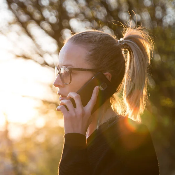 Bakgrundsbelyst bakre vy av ung kvinna talar på mobiltelefon utomhus i parken vid solnedgången. Flicka hålla mobiltelefon, med hjälp av digital enhet, titta på nedgående sol — Stockfoto