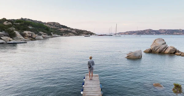 Hermosa mujer en vestido de verano de lujo de pie en el muelle de madera disfrutando de un paisaje marino tranquilo al atardecer. Vacaciones, resort y viajes . — Foto de Stock