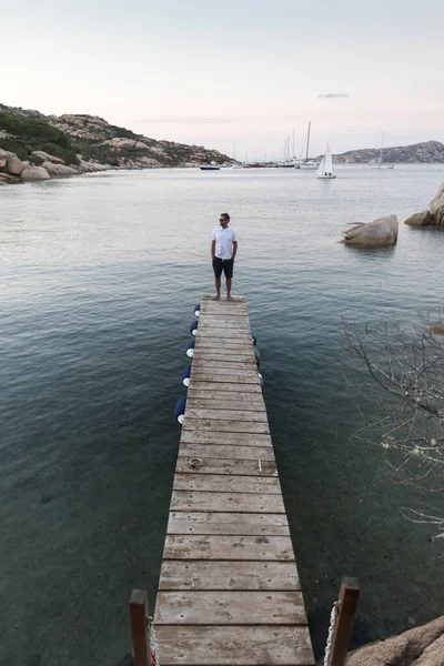 Человек, стоящий на деревянном пирсе и наслаждающийся спокойным морским пейзажем в сумерках. Мужской турист стоит на деревянном пирсе в Порто Рафаэль, Коста Смеральда, Сардиния, Италия — стоковое фото
