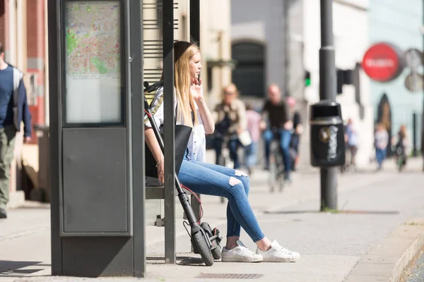 Casual caucasian teenager commuter with modern foldable urban electric scooter sitting on a bus stop bench waiting for metro city bus. Urban mobility concept — Stock Photo, Image
