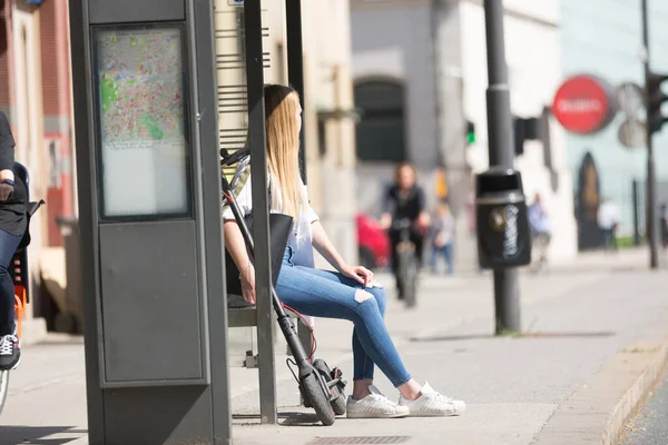 Casual kaukasiska tonåring pendlare med modern vikbar urban elektrisk skoter sitter på en busshållplats bänk väntar på tunnelbana stadsbuss. Begreppet rörlighet i städer — Stockfoto