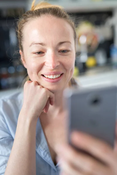 Unga leende glad nöjd kvinna inomhus i köket med hjälp av sociala medier apps på mobiltelefon för att chatta och sticka i samband med sina nära och kära. Stanna hemma, social distanserande livsstil. — Stockfoto