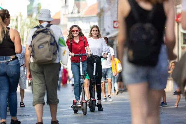 Chicas adolescentes fashinables de moda montando scooters eléctricos de alquiler público en el entorno urbano de la ciudad. Nuevo transporte público moderno ecológico en Liubliana, Eslovenia — Foto de Stock