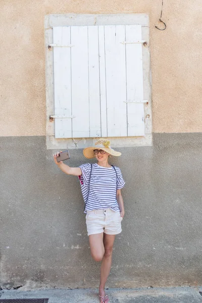 Vacker ung kvinnlig turist kvinna bär stora halmhatt, tar självporträtt selfie, står framför vita vinatage trä fönster och texturerad stenmur på gamla Medelhavet stad — Stockfoto