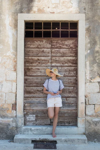 Vacker ung kvinnlig turist kvinna står framför vinatage trä dörr och strukturerad stenmur i gamla Medelhavet stad, ler, håller, smart telefon till nätverk på semestrar — Stockfoto