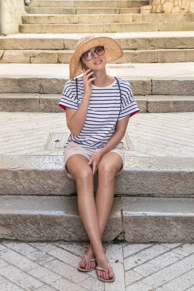 Mulher turística jovem bonita em férias de verão, sentado em degraus de pedra velhos da antiga cidade costeira medieval do Mediterrâneo, sorrindo, falando ao telefone com seus entes queridos em casa — Fotografia de Stock