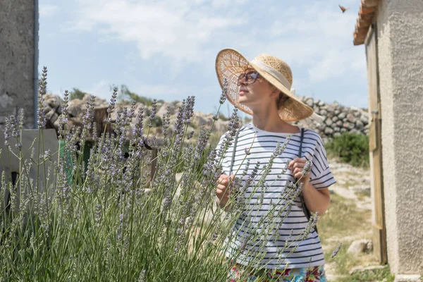 Hermosa rubia joven viajera con sombrero de sol de paja disfrutando del verano en el Mediterráneo costo pasear entre flores de lavanda en el jardín tradicional pueblo costero —  Fotos de Stock