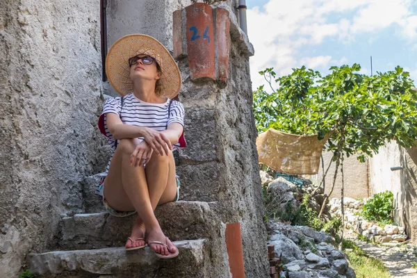 Hermosa turista femenina con sombrero de sol de paja grande y pantalones cortos sentado y relajante en las escaleras de la antigua casa de piedra durante el viaje de verano en el Mediterráneo costo en el día caluroso de verano —  Fotos de Stock