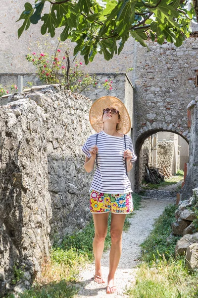 Linda loira jovem viajante vestindo chapéu de sol de palha desfrutando de verão no custo mediterrâneo, colhendo frutas sob uma figueira com flores de lavanda e casa de pedra antiga tradicional no fundo — Fotografia de Stock