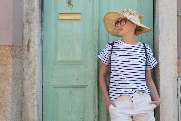 Mulher turística jovem bonita usando chapéu de sol, de pé e relaxante na sombra na frente da porta de madeira vinatage na cidade velha do Mediterrâneo, enquanto passeia no dia quente de verão — Fotografia de Stock