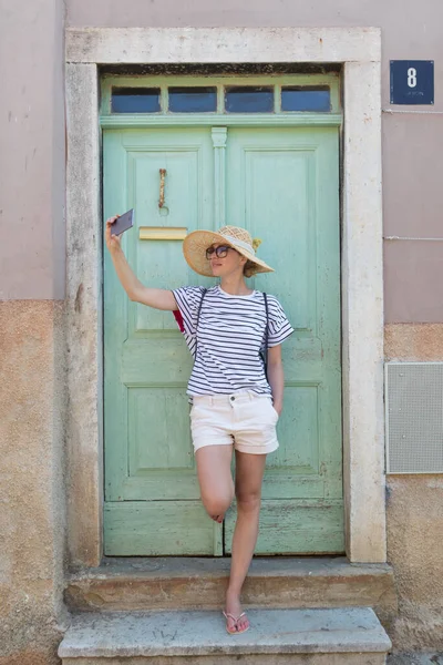 Красивая молодая женщина-туристка в большой соломенной шляпе, делает автопортрет селфи, стоя перед бирюзовой деревянной дверью виноградника и текстурированной каменной стеной в старом средиземноморском городе — стоковое фото