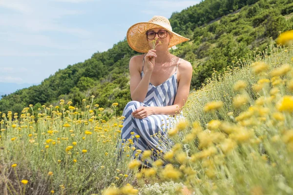 Mujer joven con vestido de verano a rayas y sombrero de paja en cuclillas en super floración de flores silvestres, relajante, recoger y oler flores en la hermosa naturaleza del mar Adriático naturaleza costera de Croacia —  Fotos de Stock