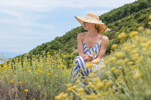 Ung kvinna bär randig sommarklänning och halmhatt sitter i super blomning av vilda blommor, koppla av samtidigt som du njuter av vacker natur Adriatiska havets kustnatur Kroatien. — Stockfoto