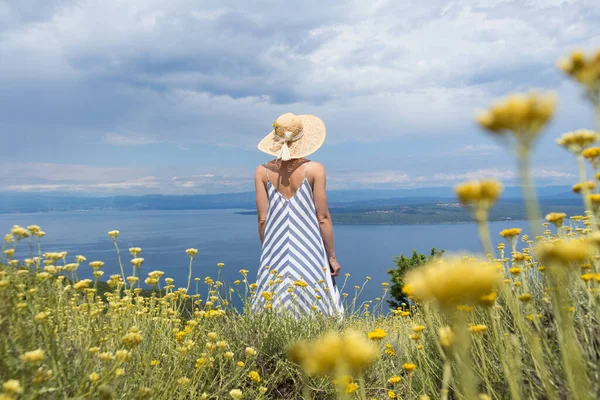 Bakifrån av ung kvinna bär randig sommar klänning och halm hatt stående i super blomning av vilda blommor, avkopplande samtidigt som den vackra utsikten över Adriatiska havet natur, Kroatien — Stockfoto