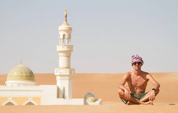 Hombre viajando en Omán, en topless usando un turbante y pantalones cortos, sentado en enviar duna en fron de mezquita en el desierto de Omán —  Fotos de Stock