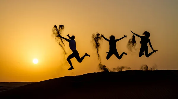 Silhouet van gelukkige reizende mensen springen op zandduin en gooien zand in de lucht in gouden zonsondergang uur — Stockfoto