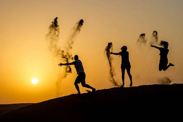 Silueta de gente feliz viajando saltando en la duna de arena y lanzando arena en el aire en la hora dorada del atardecer —  Fotos de Stock