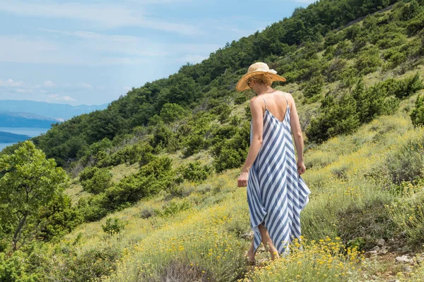 Mujer joven con vestido de verano a rayas y sombrero de paja caminando entre la floración estupenda de flores silvestres, relajante mientras disfruta de la hermosa naturaleza de la costa del mar Adriático de Croacia —  Fotos de Stock