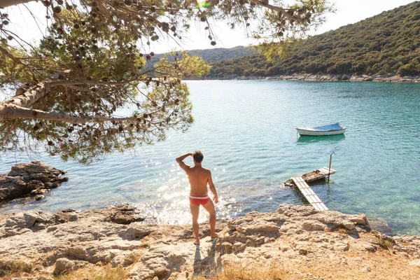 Vista trasera del hombre con speedos rojos bronceado y realaxing en la cala salvaje del mar Adriático en una playa a la sombra de pino. — Foto de Stock