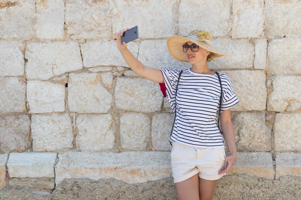 Vacker ung kvinnlig turist kvinna bär stora halmhatt, tar självporträtt selfie, står framför gamla texturerade stenmur i gamla Medelhavet stad — Stockfoto