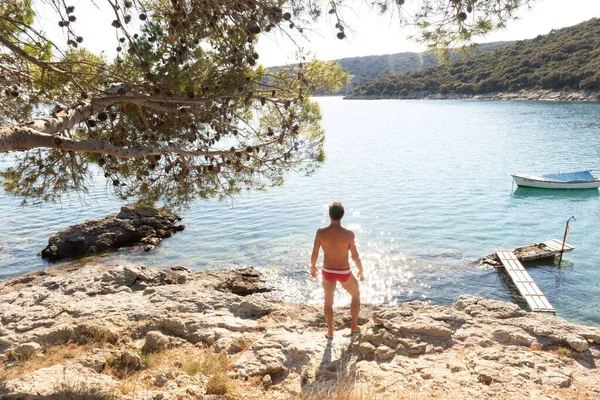 Vista trasera del hombre con speedos rojos bronceado y realaxing en la cala salvaje del mar Adriático en una playa a la sombra de pino. — Foto de Stock