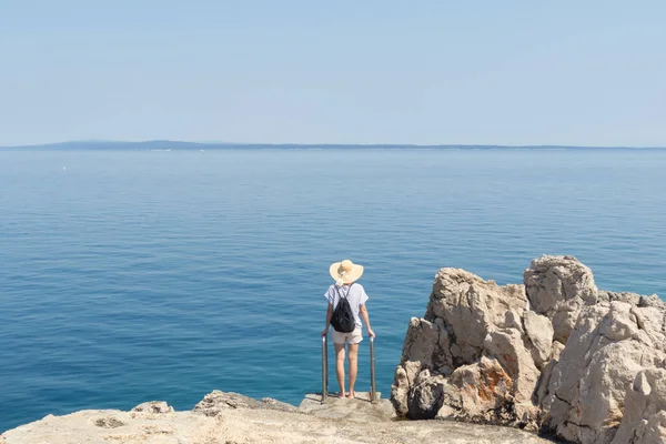 穿着稻草夏帽和背包的女旅行家站在岩石悬崖的边缘，凝视着地平线上蓝色的大海和岛屿 — 图库照片