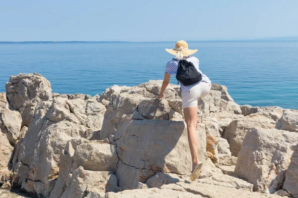 女旅行家头戴稻草夏帽，背着背包，爬上悬崖边，望着蓝色的大海和地平线上的岛屿 — 图库照片