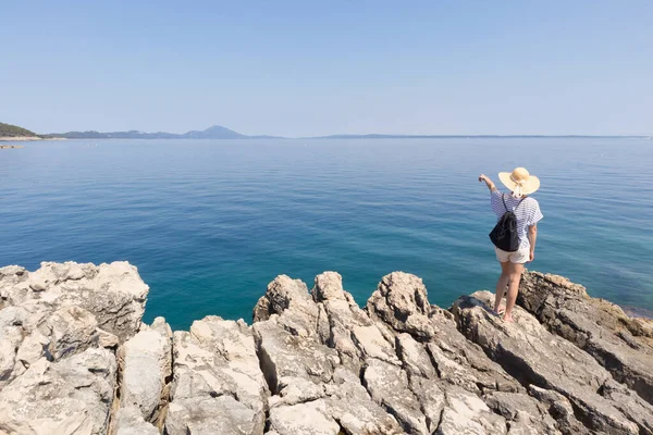 Donna viaggiatore indossa cappello estivo di paglia e zaino, in piedi sul bordo della scogliera rocciosa guardando e indicando il grande mare blu e le isole all'orizzonte — Foto Stock