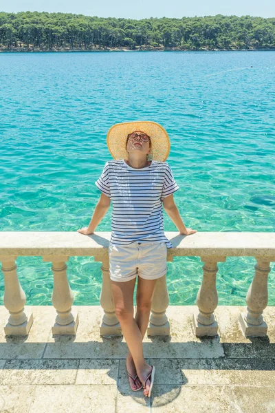 Visão traseira da mulher usando chapéu de verão de palha, inclinando-se contra a cerca de pedra velha elegante da vila costeira, relaxante enquanto olha para o mar Adriático azul, na ilha de Losinj Croácia. — Fotografia de Stock