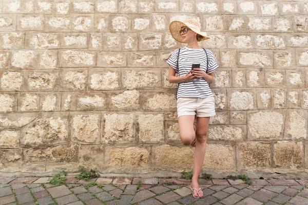 Красивая молодая женщина-туристка стоит перед старой текстурированной каменной стеной в старом средиземноморском городе, улыбаясь, держа, смартфон к сети на vacationes. Копирование пространства — стоковое фото