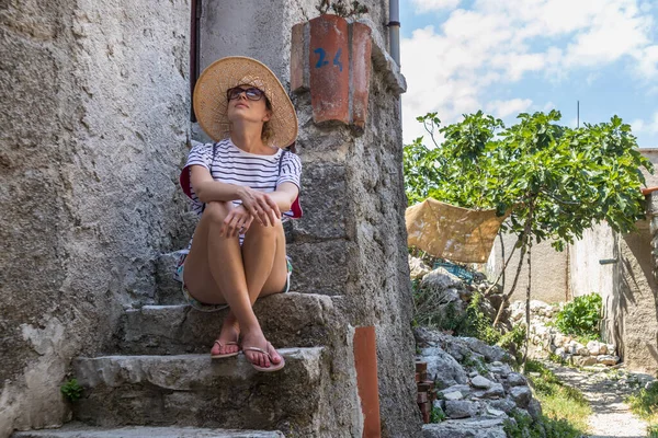 Linda turista feminina vestindo grande chapéu de sol palha e shorts sentado e relaxante em escadas casa de pedra velha durante as viagens de verão no custo Mediterrâneo no dia quente de verão — Fotografia de Stock