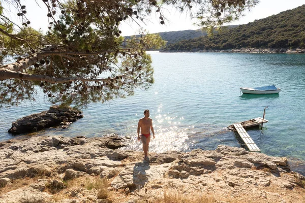 Joven deportista con speedos rojos bronceado y realaxing en la cala salvaje del mar Adriático en una playa a la sombra de pino — Foto de Stock