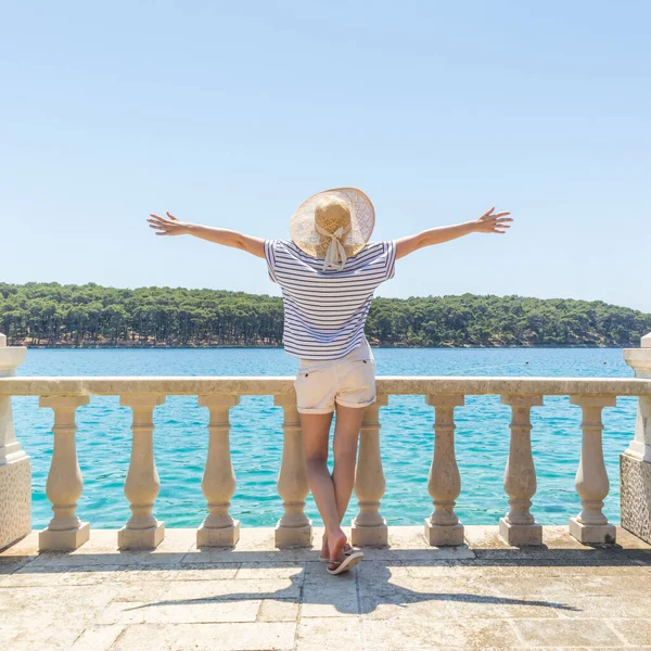 Vista trasera de la mujer feliz de vacaciones, con sombrero de verano de paja, de pie en el elegante y lujoso balcón de piedra antigua de la villa costera, relajante, los brazos levantados al sol, mirando al mar Adriático azul —  Fotos de Stock