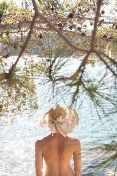 Rückansicht der oben ohne schönen Frau, die nichts als einen Strohhut trägt und sich an der wilden Küste der Adria an einem Strand im Schatten der Pinien erholt. — Stockfoto